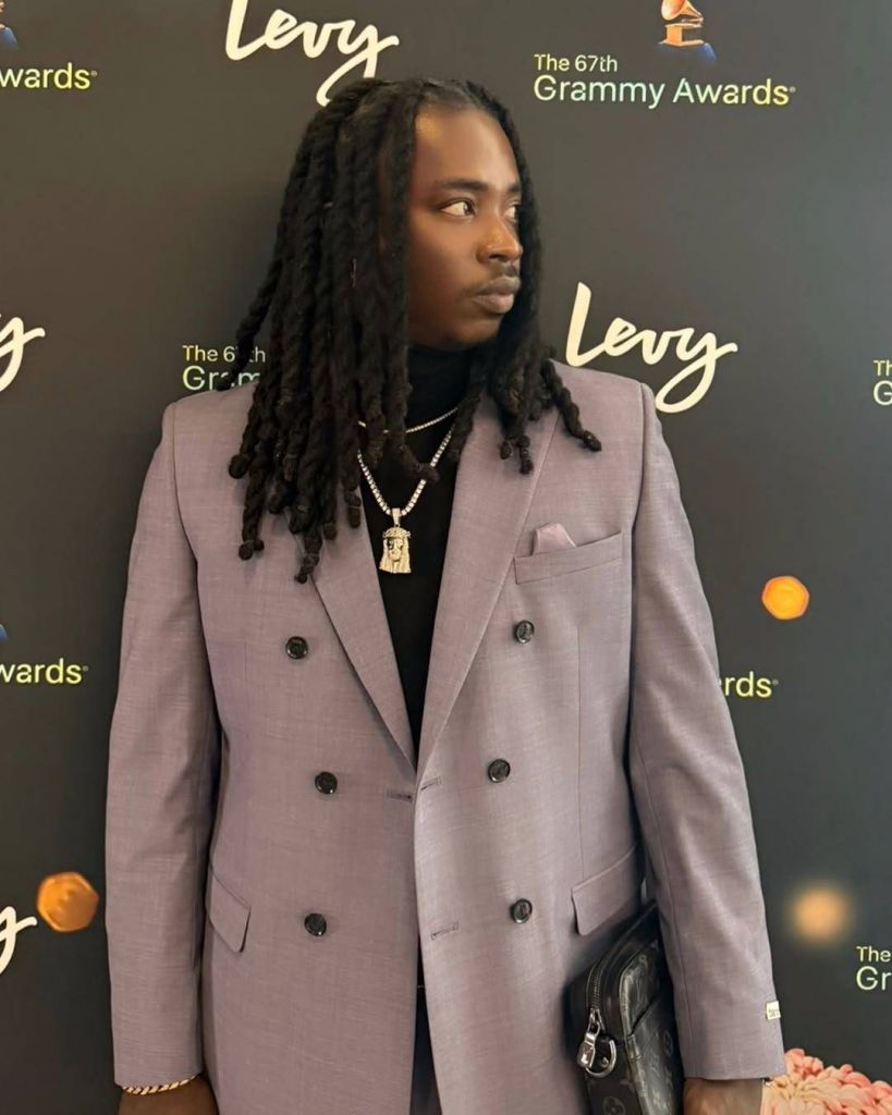 Nigerian skitmaker Lord Lamba stands confidently on a red carpet at the 66th Grammy Awards, dressed in a stylish double-breasted gray suit over a black shirt. His long, twisted braids cascade over his shoulders, complemented by a gold chain with a prominent pendant and a gold bracelet on his wrist. He holds a sleek black designer bag, while a pocket square adds a touch of elegance to his look. The backdrop features the Levy Awards and Grammy Awards logos, highlighting the prestigious event.