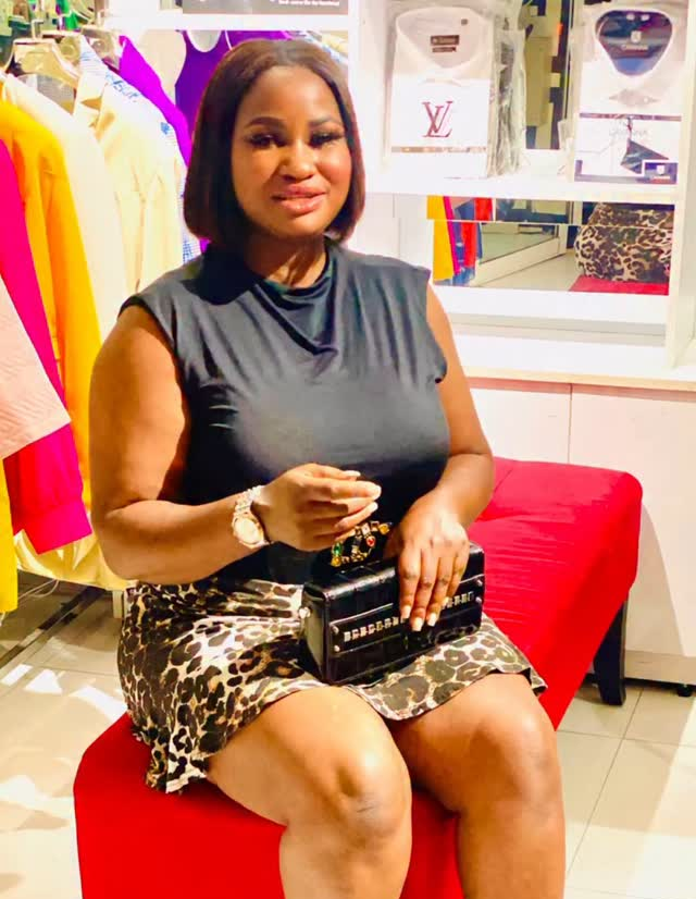 The scene features Hellen Ati, the alleged Kenyan baby mama of Cubana Chief Priest, seated on a red bench in a vibrant clothing store. She wears a sleeveless dark top paired with a leopard-print skirt, accessorized with a stylish black handbag adorned with embellishments and a wristwatch. Behind her, the store displays colorful garments, including a prominent Louis Vuitton shirt, and a mirror reflects part of the setting, adding depth to the stylish environment.