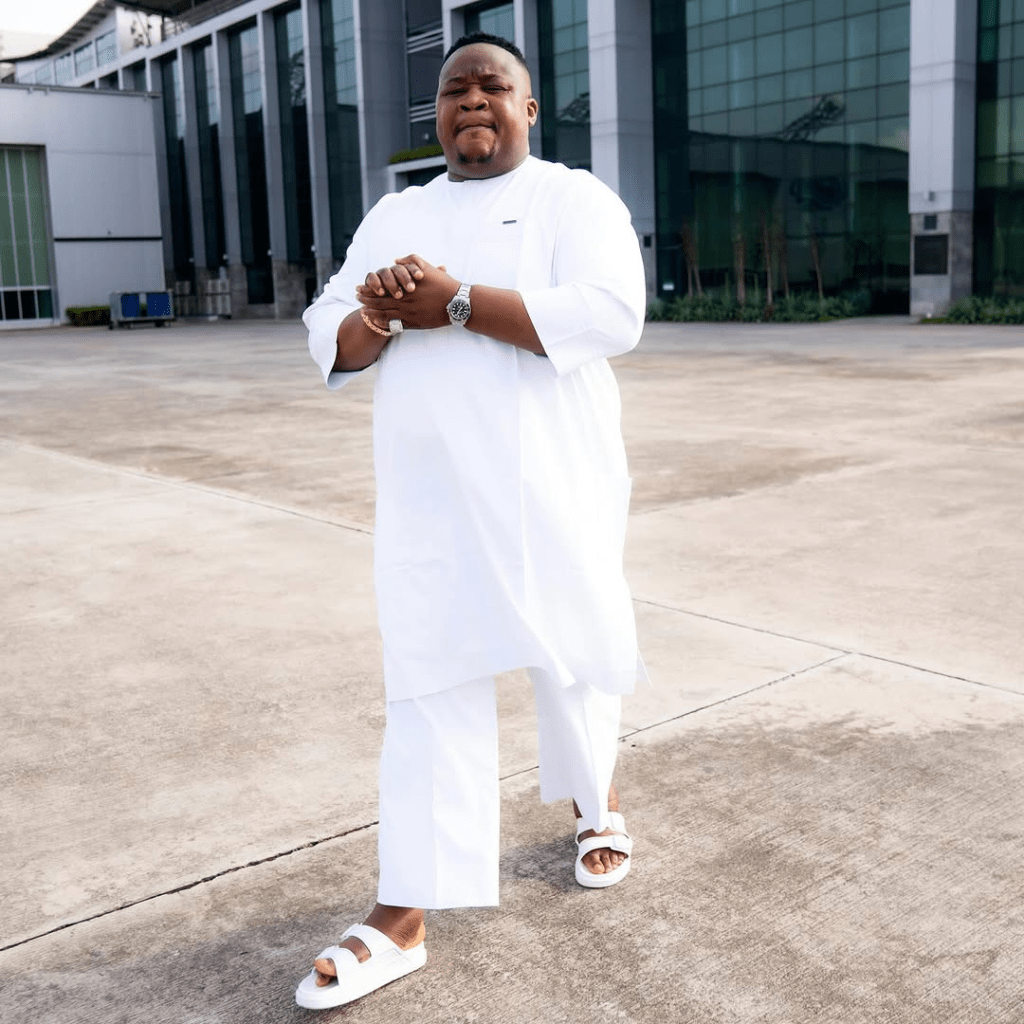 Cubana Chief Priest stands confidently on a concrete pavement in front of a modern glass building, dressed in an all-white traditional outfit with a subtle logo detail. His arms are crossed, showcasing a wristwatch and a gold bracelet, while he wears white sandals. The early morning light highlights the sleek architecture and surrounding greenery, creating a stylish urban backdrop.