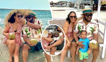 Businessman Olakunle Churchill sits on a bench with his wife and young son at a sandy beach setting, under a white canopy with a colorful fabric backdrop. Churchill wears a patterned shirt, dark shorts, a green-striped straw hat, and "ICEBERG" sandals, holding a coconut drink. His wife, in a red floral dress and sunglasses, sits barefoot with a woven hat and basket beside her. Their son, dressed in a blue rash guard and yellow shorts with "KING" text, stands between them, holding a coconut. The background features a cloudy sky, metal structures, and distant beach activity.