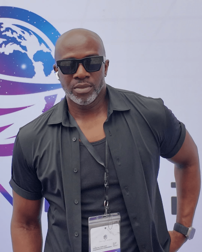 Toke Makinwa's ex-husband, Maje Ayida, stands confidently in a dark short-sleeved shirt over a black top, accessorized with sunglasses and a smartwatch. He wears a badge with a lanyard, set against a backdrop featuring a stylized world map and vibrant purple and blue hues.