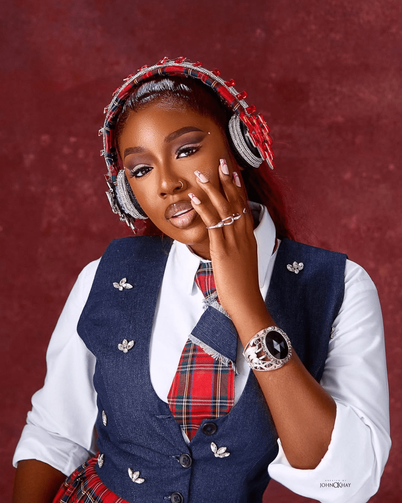 BBNaija star Tolanibaj poses in a stylish school uniform featuring a blue denim vest with decorative pins, a red plaid tie, and a white shirt, accessorized with a jeweled headband, headphones, and a statement watch, set against a rich red background.