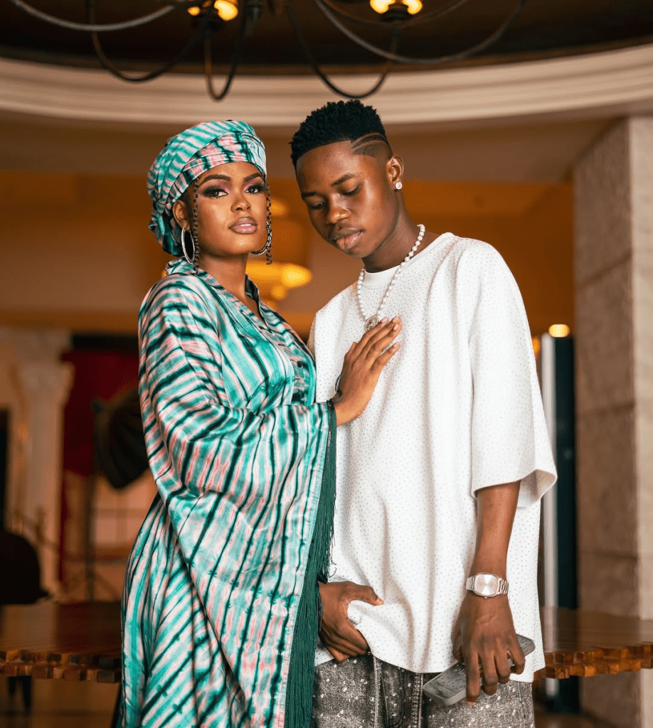 Tiktoker Peller and his girlfriend Jarvis stand closely together in an elegant indoor setting with warm lighting and a chandelier overhead. Peller wears a white outfit with a pearl necklace and a watch, while Jarvis is dressed in a vibrant, tie-dye robe with a matching headwrap, accessorized with large earrings.