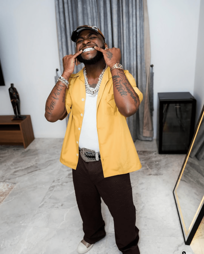 Singer Davido, wearing a yellow shirt, white undershirt, and dark pants with a large belt buckle, accessorizes with multiple chains, bracelets, and a cap, while standing in a modern room with a mirror, furniture, and marble floor.