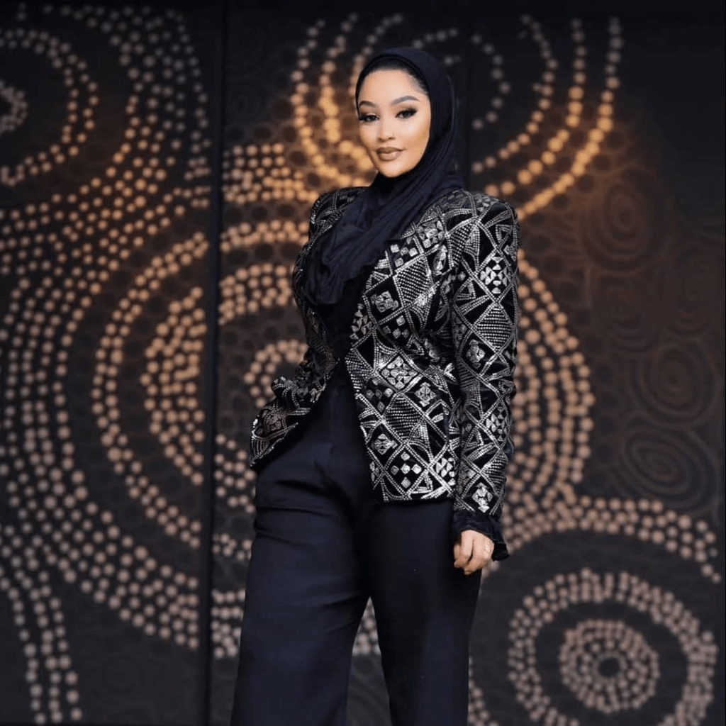 Zari, ex-girlfriend of singer Diamond Platnumz, stands confidently in an elegant black outfit with a shimmering, intricately patterned jacket and matching pants, set against a stylish backdrop with circular designs.