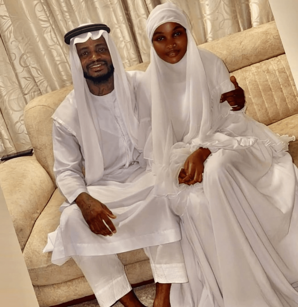Singer Diamond Platnumz and his new wife Zuchu sit together on a beige couch, both dressed in elegant white traditional attire, with intricate headwear and flowing garments, set against a softly lit backdrop with patterned curtains.