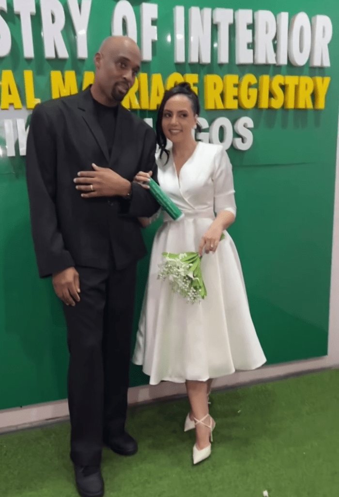 Businessman Dilly and his newly married wife Anastasia pose together in front of a sign reading "MINISTRY OF INTERIOR NATIONAL MARRIAGE REGISTRY LAGOS," with Anastasia holding a bouquet of white flowers.