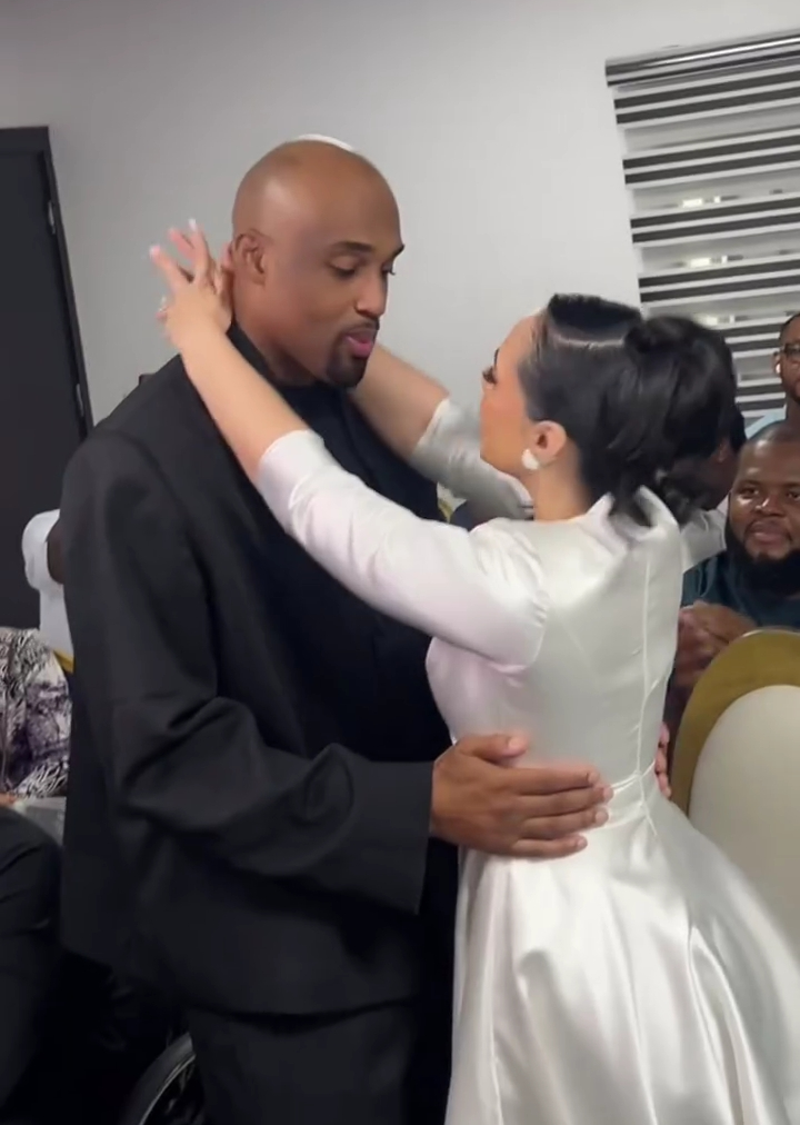 Businessman Dilly and his newly married wife Anastasia share a close embrace during a ceremony, with guests seated in the background and a striped window covering visible.