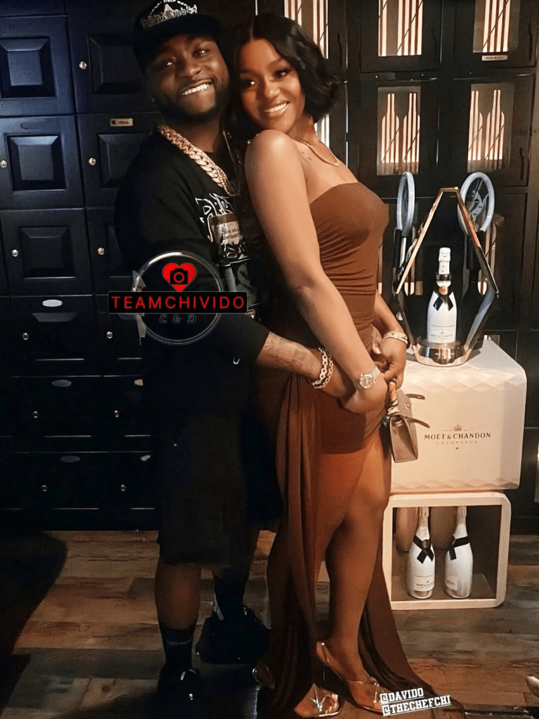 Singer Davido and his wife Chioma pose affectionately, with Davido in a black outfit and cap, adorned with a gold chain, and Chioma in a chic brown dress, standing beside a Moët & Chandon champagne display in a stylish, dimly lit setting with dark cabinetry.