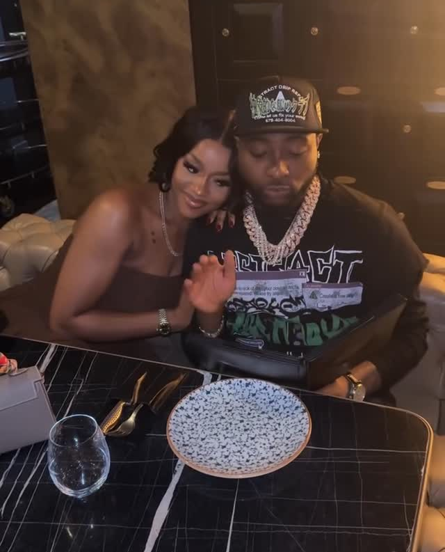 Singer Davido and his wife Chioma are seated closely at a stylish dining table with a marble top, featuring a decorative plate, wine glass, and cutlery. Davido wears a black shirt with bold text and a cap, accessorized with a chunky chain, while Chioma, in a strapless top, rests her hand on his shoulder, both exuding a relaxed and affectionate vibe in a warmly lit setting.