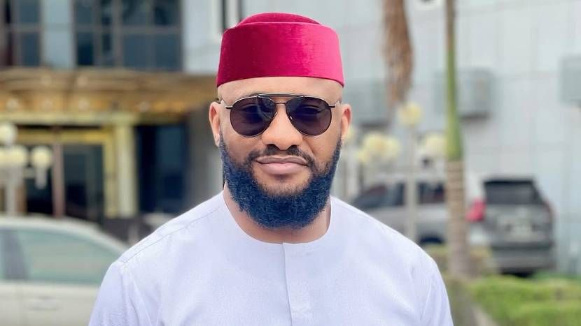 Actor Yul Edochie stands outdoors in front of a building, wearing a white traditional outfit and a red cap, with sunglasses and a well-groomed beard, as cars and greenery are visible in the background.