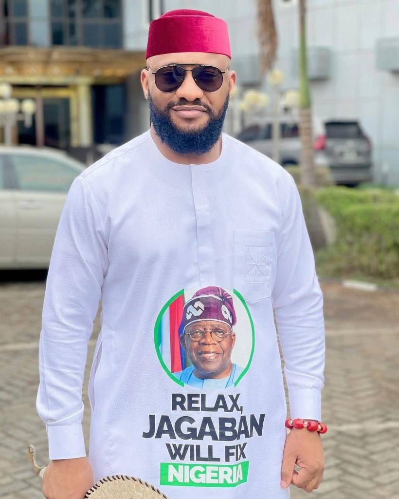 Actor Yul Edochie stands outdoors wearing a white long-sleeve shirt featuring a portrait of a man in a purple cap, with the text "RELAX, JAGABAN WILL FIX NIGERIA" printed below. He accessorizes with a red cap, sunglasses, a red beaded bracelet, and holds a straw hat in one hand.