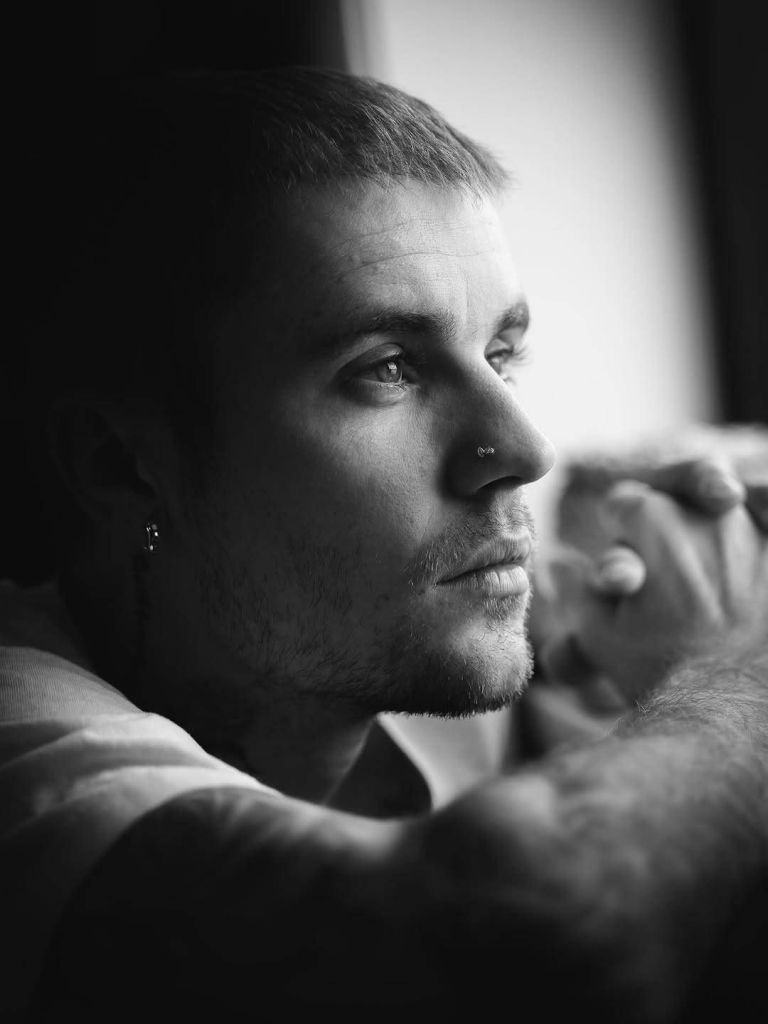 A black-and-white close-up of popstar Justin Bieber with short hair, a small nose piercing, and an earring, gazing thoughtfully out a window while resting his arm.