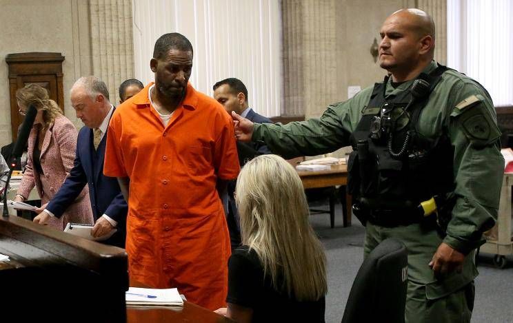 R. Kelly in an orange prison jumpsuit being escorted by a guard in a courtroom, with several people seated and standing around a wooden table.