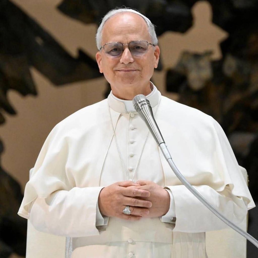 Pope Leo XIV in white papal attire, standing with hands clasped, speaking at a microphone with a blurred background.