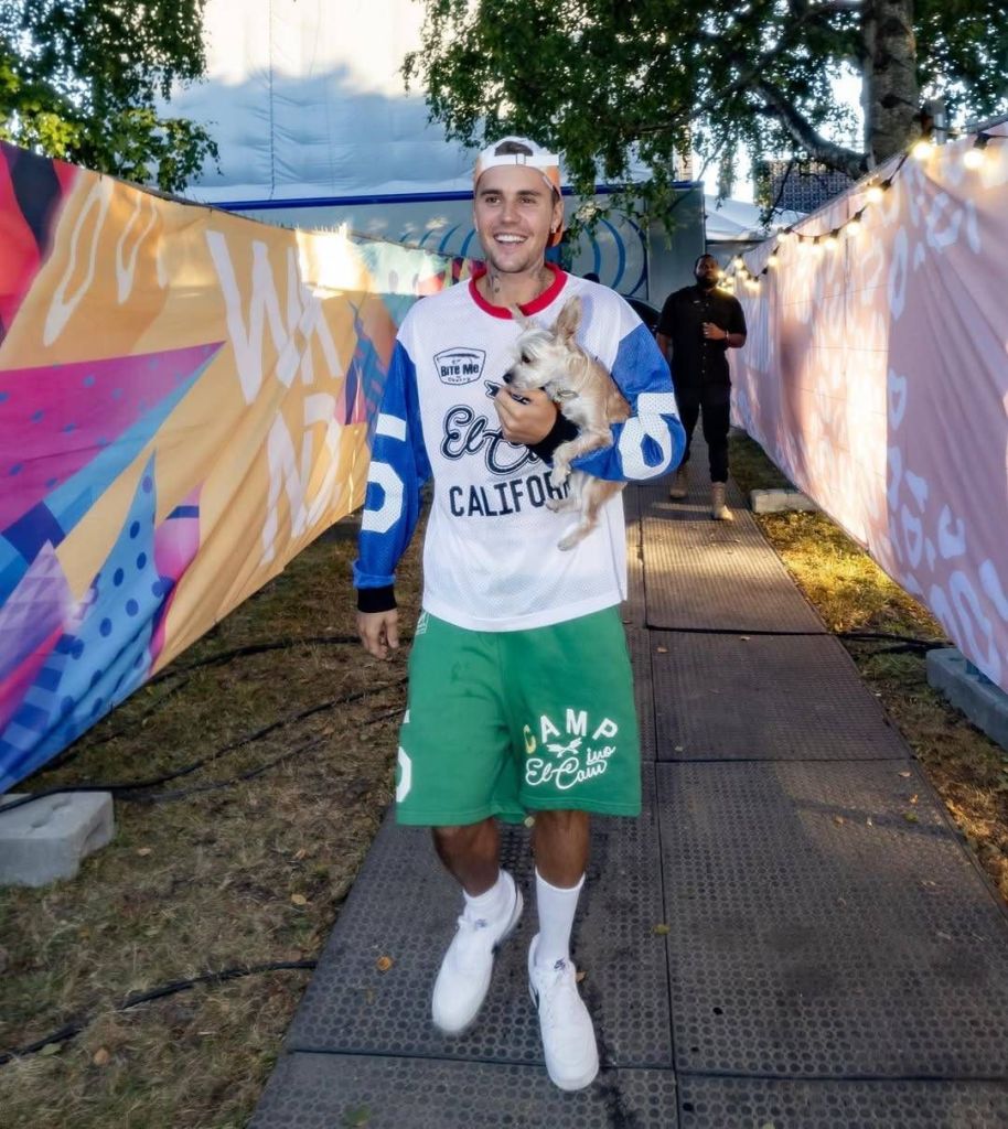Justin Bieber wearing a "Bite Me California" jersey, green "Camp Flog Gnaw" shorts, and white sneakers, holding a small dog while walking on a pathway lined with colorful banners and string lights.