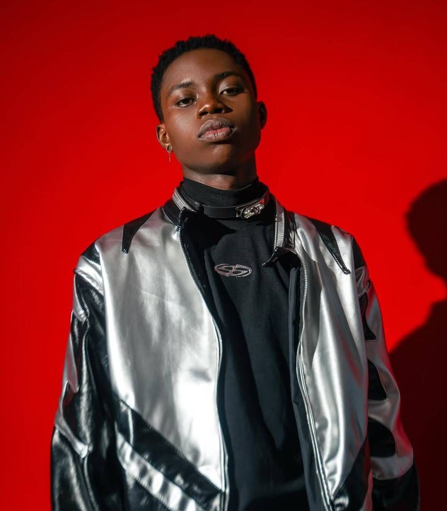 Tiktoker Peller stands against a vibrant red background, wearing a striking silver and black jacket over a black shirt, accessorized with a choker and earrings.