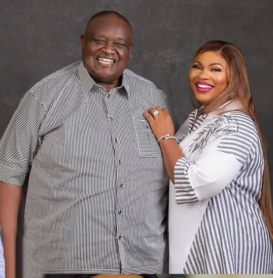 Late Chief (Engr.) Emmanuel Iwuanyanwu stands smiling in a striped shirt, with his widow Frances Chinonyerem Enwerem beside him, also smiling, wearing a matching striped and white outfit with delicate embellishments.