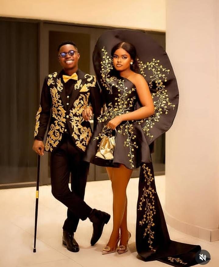 Tiktoker Peller and his girlfriend Jarvis stand together in elegant attire. Peller sports a black suit with intricate gold embroidery and a bow tie, holding a cane, while Jarvis wears a striking black dress with gold floral accents and a flowing train, accessorized with a gold clutch and heels.