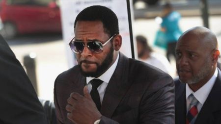 Singer R. Kelly wearing sunglasses and a dark suit, gesturing with his hand, sits next to an older man in a suit at an outdoor event with a red car and people in the background.