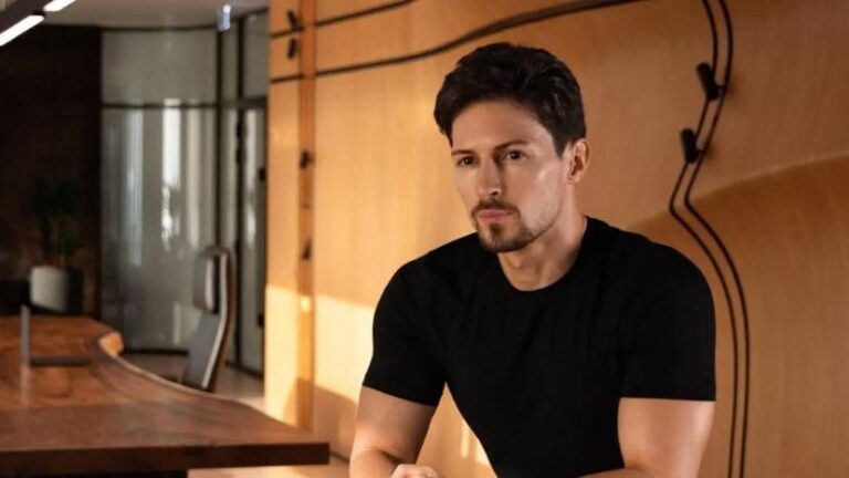 Telegram founder Pavel Durov sits in a modern office with wooden walls and a curved desk, wearing a black t-shirt, with natural light illuminating the room.