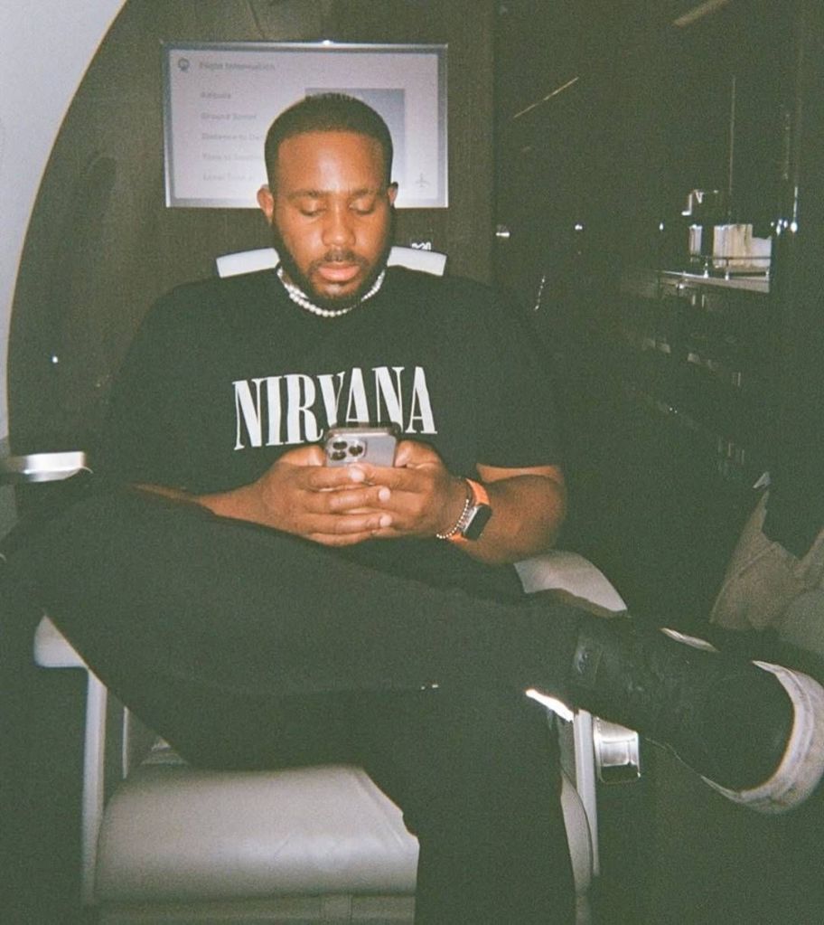 BBNaija star Kiddwaya sits inside a private jet, wearing a black Nirvana t-shirt and pearl necklace, engrossed in his phone, with a flight information screen in the background.