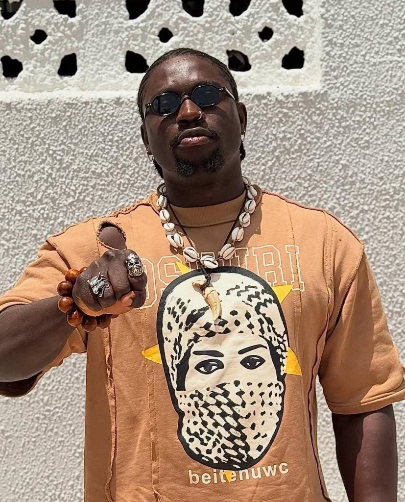 Activist VeryDarkMan stands outdoors against a textured wall, wearing a distressed orange shirt with a patterned face design, accessorized with multiple shell necklaces, wooden beaded bracelets, and bold rings, pointing forward with confidence.