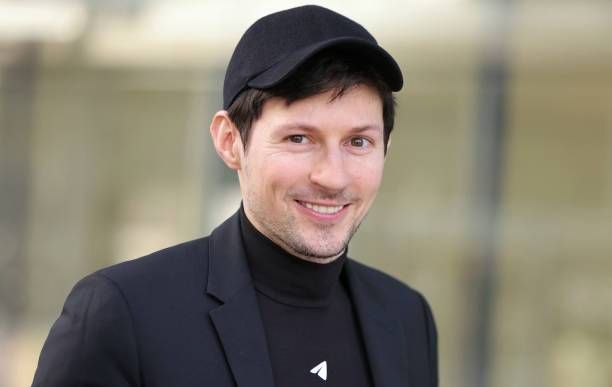 Telegram CEO Pavel Durov smiling, wearing a black cap, blazer, and turtleneck with a small logo, standing against a blurred outdoor background.