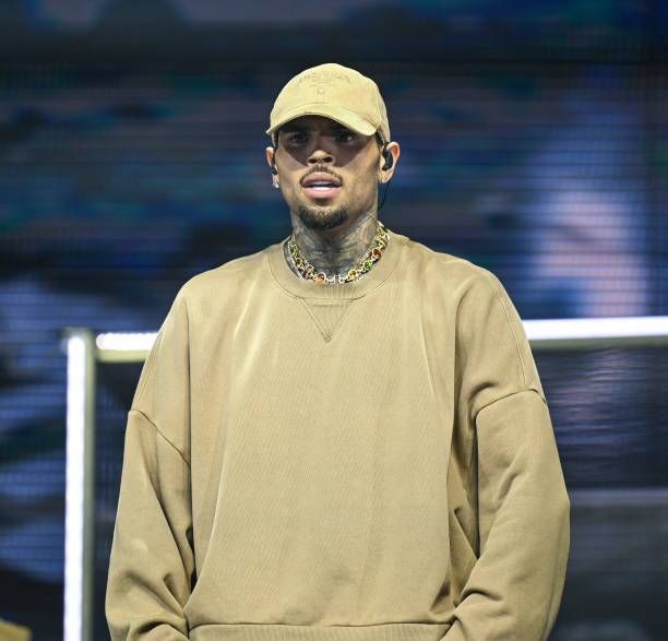 Singer Chris Brown standing on stage, wearing a beige cap and oversized sweater with a colorful chain necklace, against a vibrant background.