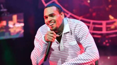 Singer Chris Brown performing on stage, holding a microphone, wearing a striped shirt, with vibrant stage lighting in the background.