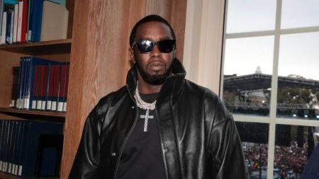 Diddy stands indoors near a bookshelf, wearing a black leather jacket, black shirt, and a large cross necklace, with sunglasses and a crowd visible through a window behind him.