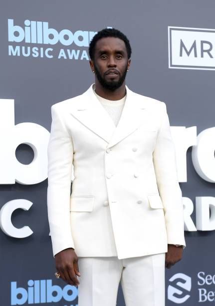 Diddy stands on the Billboard Music Awards red carpet, dressed in a sharp white double-breasted suit with a matching shirt, exuding style and confidence against the event's backdrop.