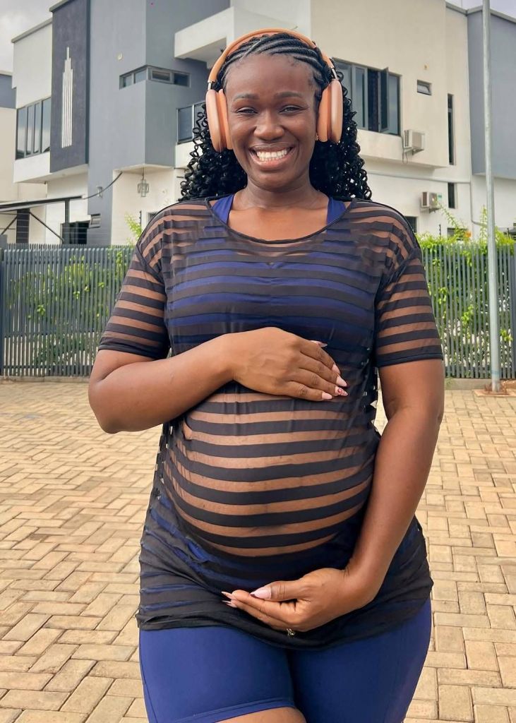 Comedienne Real Warri Pikin stands outdoors in front of modern buildings, wearing a striped sheer top and blue shorts, with her hands gently cradling her pregnant belly, smiling brightly while wearing headphones.