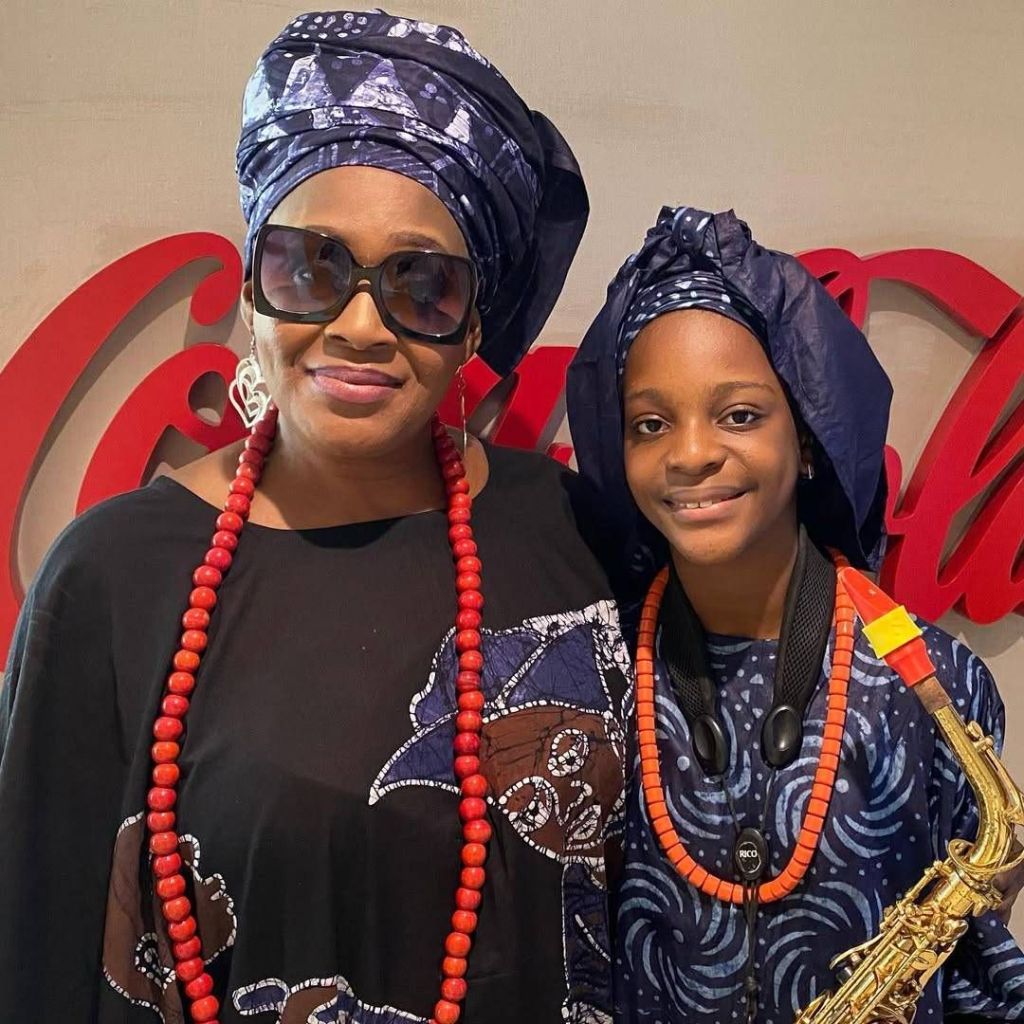 Veteran journalist Kemi Olunloyo stands alongside a young person, both adorned in traditional attire with blue headwraps and red beaded necklaces, in front of a Coca-Cola sign, with the young person holding a saxophone.