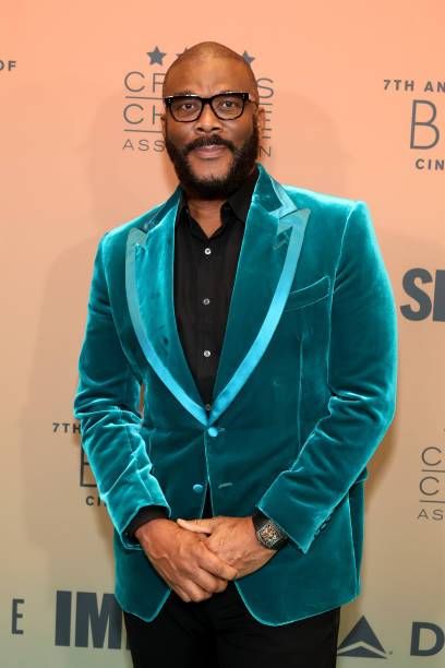 Filmmaker Tyler Perry stands on a red carpet in a teal velvet suit with a black shirt, hands clasped, against a backdrop of the Critics Choice Association and 7th Annual Black Cinema event.