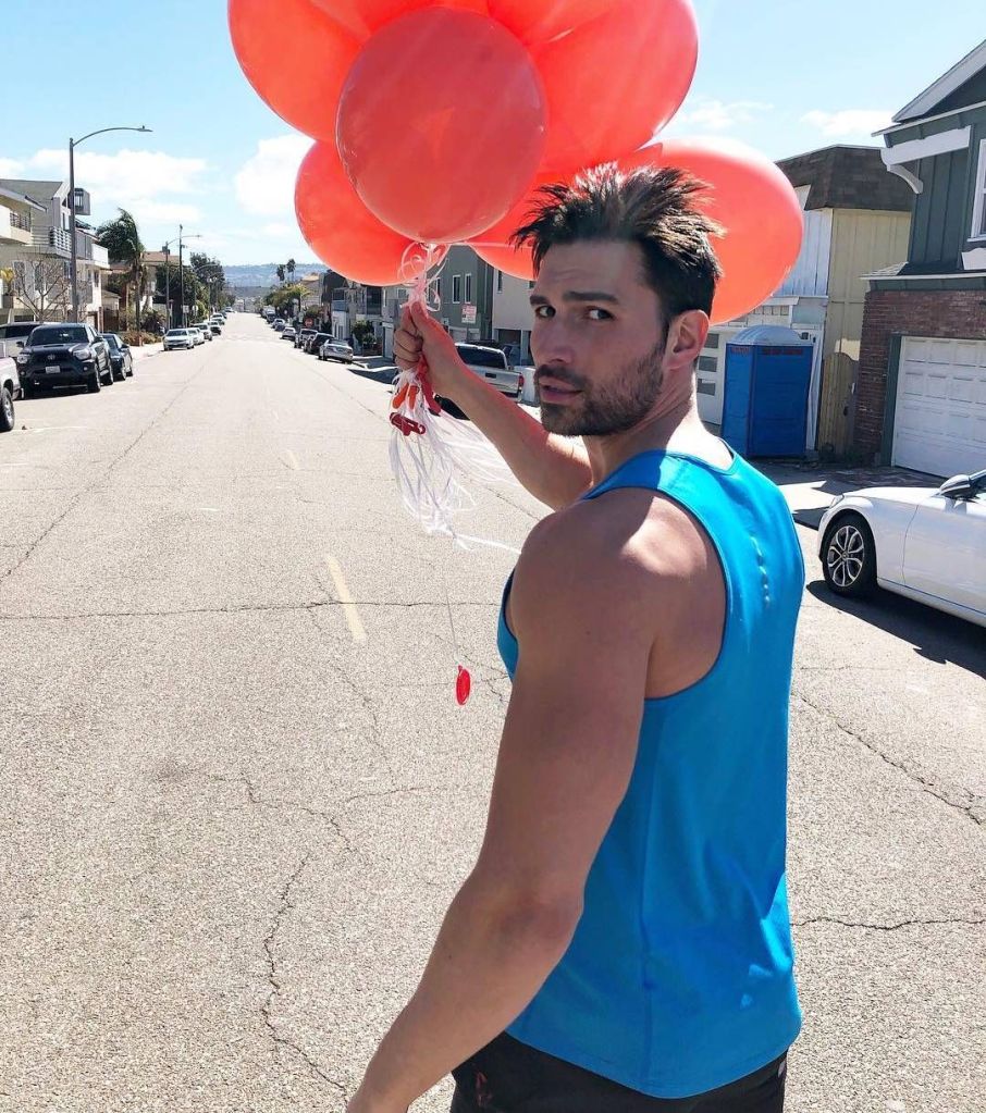 Actor Derek Dixon stands on a sunny street holding a bunch of red balloons, wearing a blue tank top, with parked cars and houses lining the road.