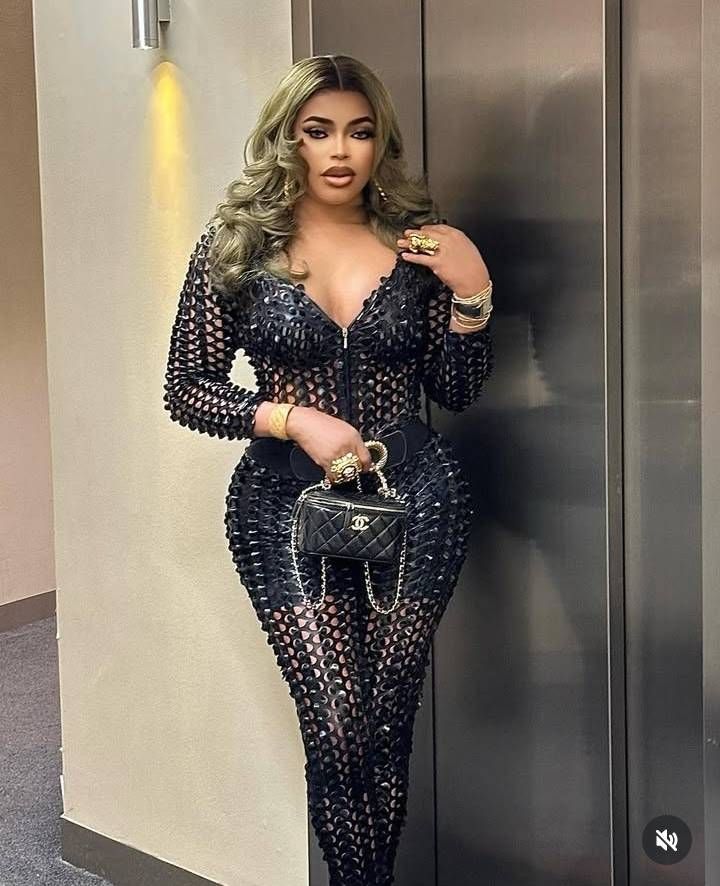 Crossdresser Bobrisky stands in an elevator, wearing a stylish black mesh bodysuit with cut-out details, accessorized with gold jewelry, a Chanel handbag, and a long, wavy blonde wig.