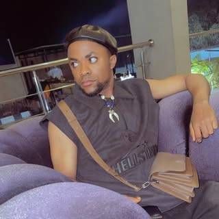 Ezeani Valentine is seated on a purple couch, wearing a sleeveless dark shirt with a "HELLSTAR" logo, a black cap, a necklace with a horn pendant, and a brown crossbody bag, with a modern indoor setting in the background.