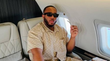 BBNaija star Kiddwaya lounges in a luxurious private jet, wearing a stylish beige outfit and sunglasses, while enjoying an elaborate meal served on multiple plates, with plush leather seats and a window view in the background.