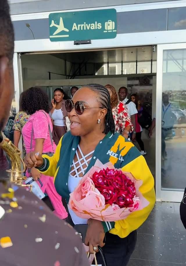 Blessing CEO is warmly welcomed at Asaba airport prior to her wedding, sporting a green and yellow "MEET" jacket and sunglasses. She holds a bouquet of red roses, surrounded by well-wishers and confetti under the "Arrivals" sign.