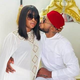 Blessing CEO shares a tender moment with her partner IVD, both dressed in elegant white outfits, as he kisses her cheek while wearing a red cap, set against a luxurious golden backdrop.