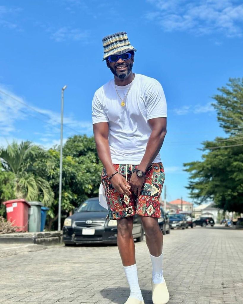 Music executive Teebillz stands on a paved street under a clear blue sky, wearing a striped bucket hat, blue sunglasses, a white shirt, colorful patterned shorts, white socks, and beige shoes, with cars and greenery in the background.