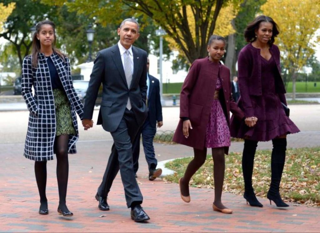 Barack Obama walks hand-in-hand with Michelle Obama and their two daughters on a brick path lined with autumn trees, all dressed in stylish coats and dresses.