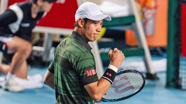 Kei Nishikori celebrates with a clenched fist, wearing a green shirt with black stripes, a white cap, and wristbands, framed by the backdrop of a tennis court and equipment.