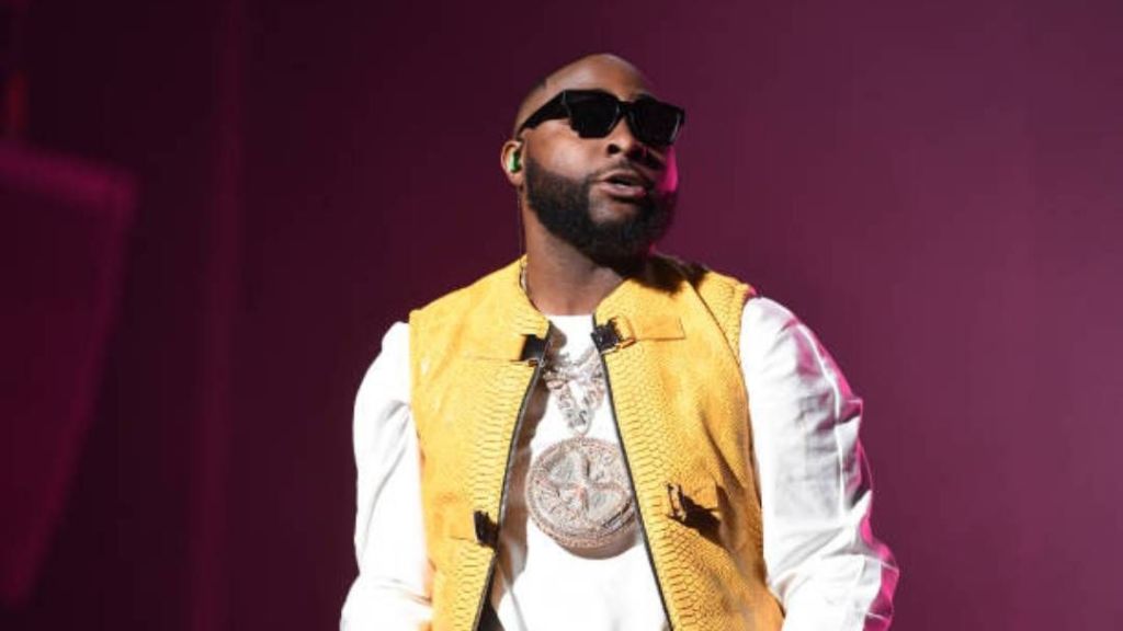 Singer Davido wears a yellow vest over a white shirt, accented by a bold, round pendant necklace, with a dark backdrop enhancing his stylish ensemble.