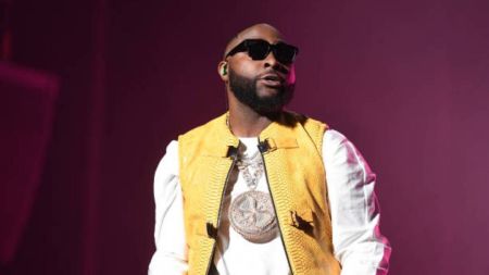 Singer Davido wears a yellow vest over a white shirt, accented by a bold, round pendant necklace, with a dark backdrop enhancing his stylish ensemble.
