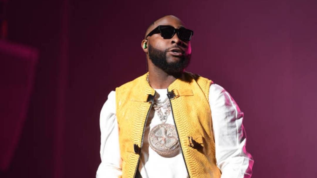 Singer Davido wears a yellow vest over a white shirt, accented by a bold, round pendant necklace, with a dark backdrop enhancing his stylish ensemble.
