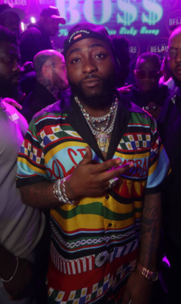 Davido sports a vibrant, patterned shirt paired with layered necklaces and bracelets, standing against a glowing neon backdrop that reads “BOSS,” evoking a high-energy party vibe.