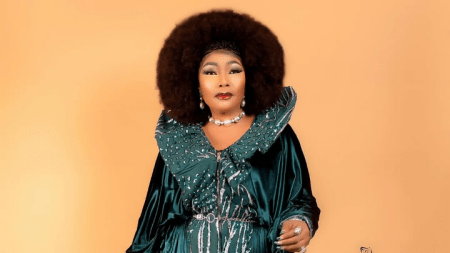 Eucharia Anunobi stands poised in a flowing green ensemble with silver embellishments, featuring a ruffled collar and belted waist. She wears a pearl necklace and a ring, set against a warm peach-toned backdrop.
