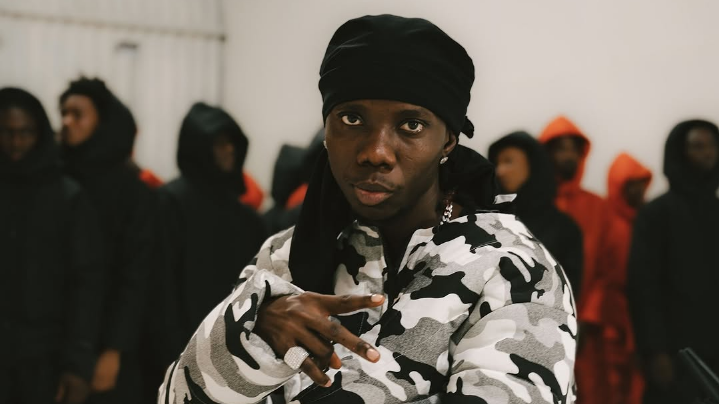 Blaqbonez, dressed in a camouflage jacket and black head covering, stands confidently indoors while making a hand gesture, flanked by masked figures in dark and red hooded outfits that create a bold visual contrast.